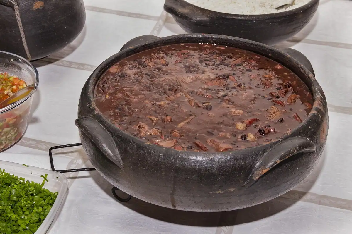 Oală neagră de lut cu fasole gătită lent, lângă ierburi proaspete tocate