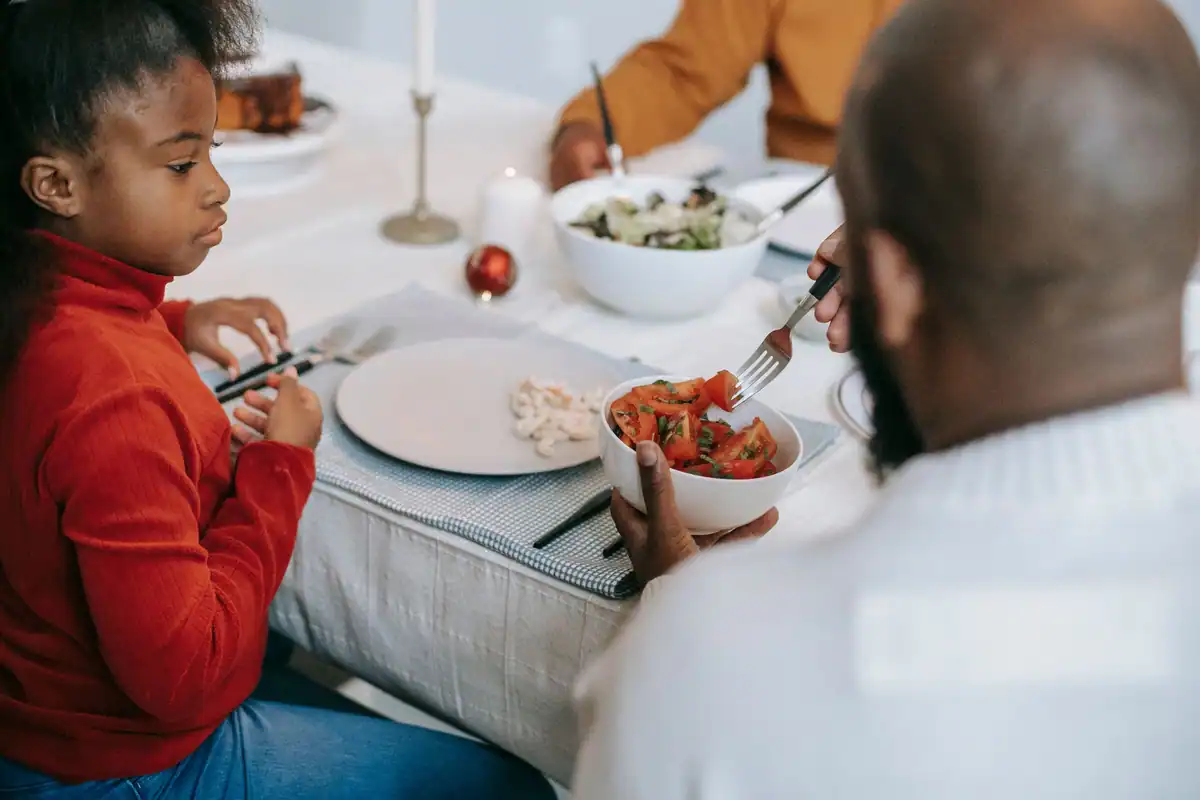 Fetiță care așteaptă la masă salata din farfurie, lângă un adult care servește
